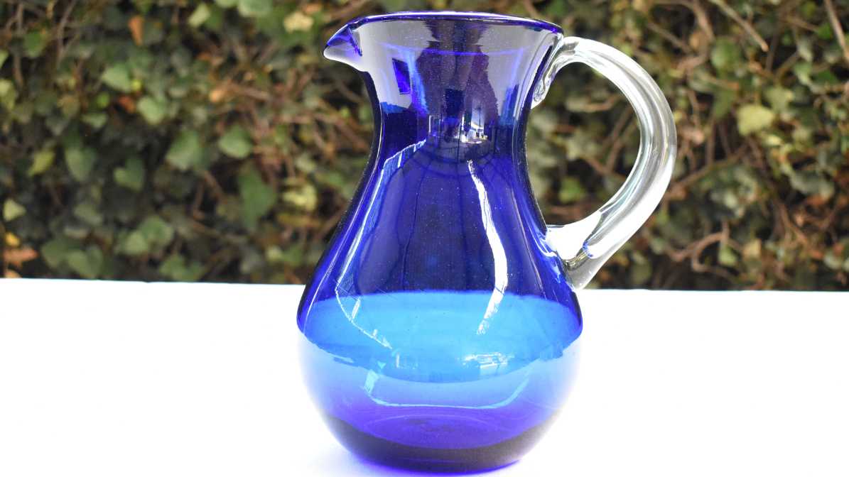 Jarra de vidrio soplado azul cobalto para agua sobre mesa con mantel blanco con fondo de hiedras sobre un muro.